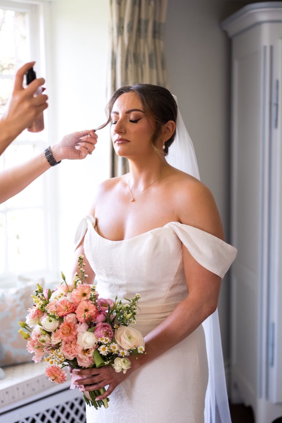 Bride with pink bouquet having her makeup finished by the window